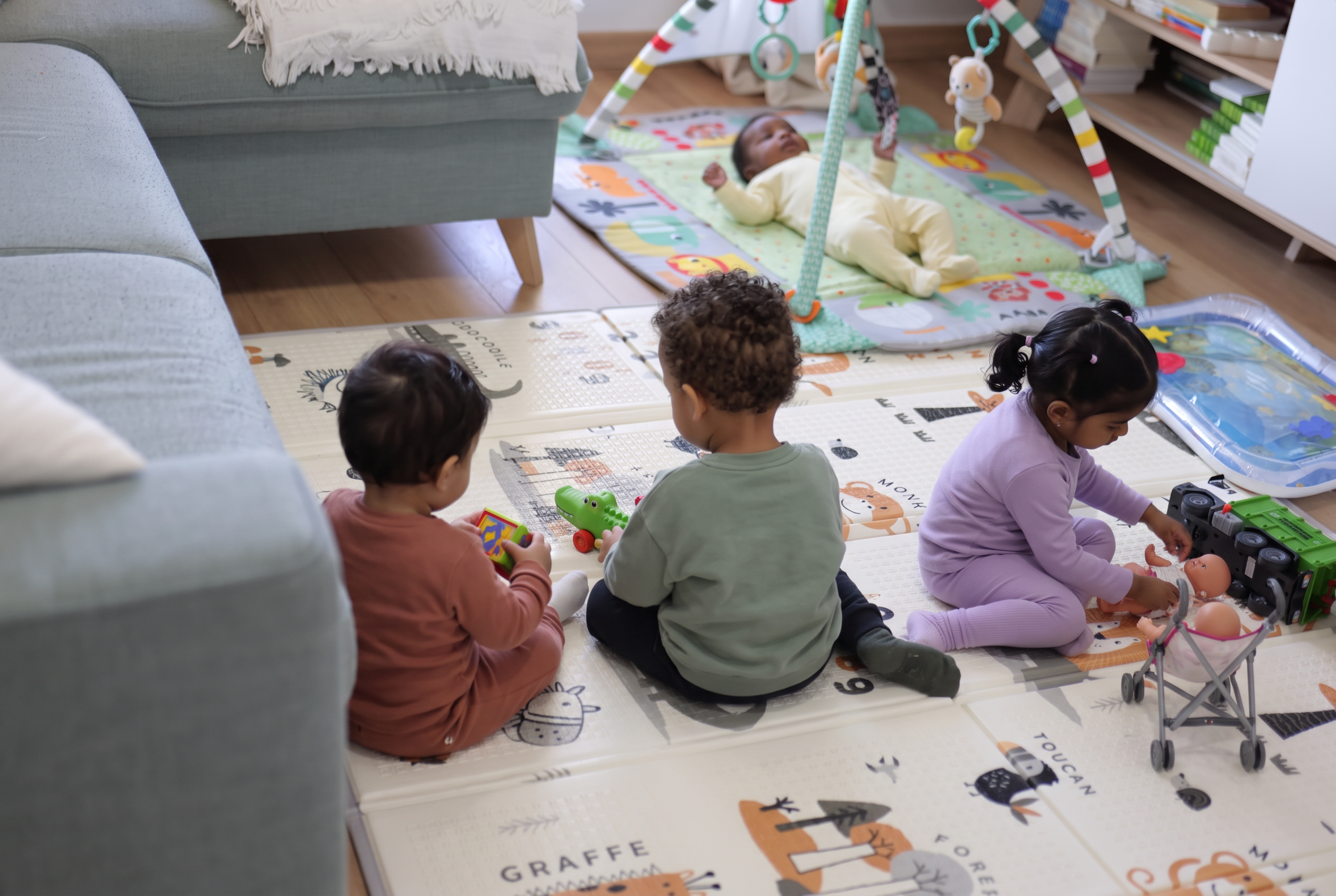 Un nourrisson sur le tapis vert et trois jeunes enfants jouant sur le tapis beige avec des jouets, une poupée et une poussette